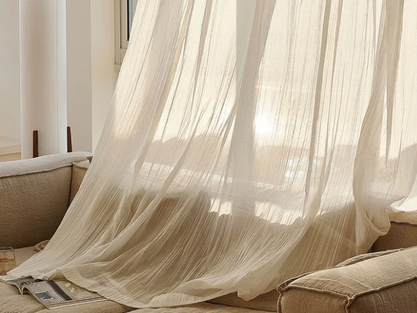 Flowing, light-filtering semi sheer curtains in a cream color, with a slight crinkle texture, draping over a linen sofa in a sunlit room.