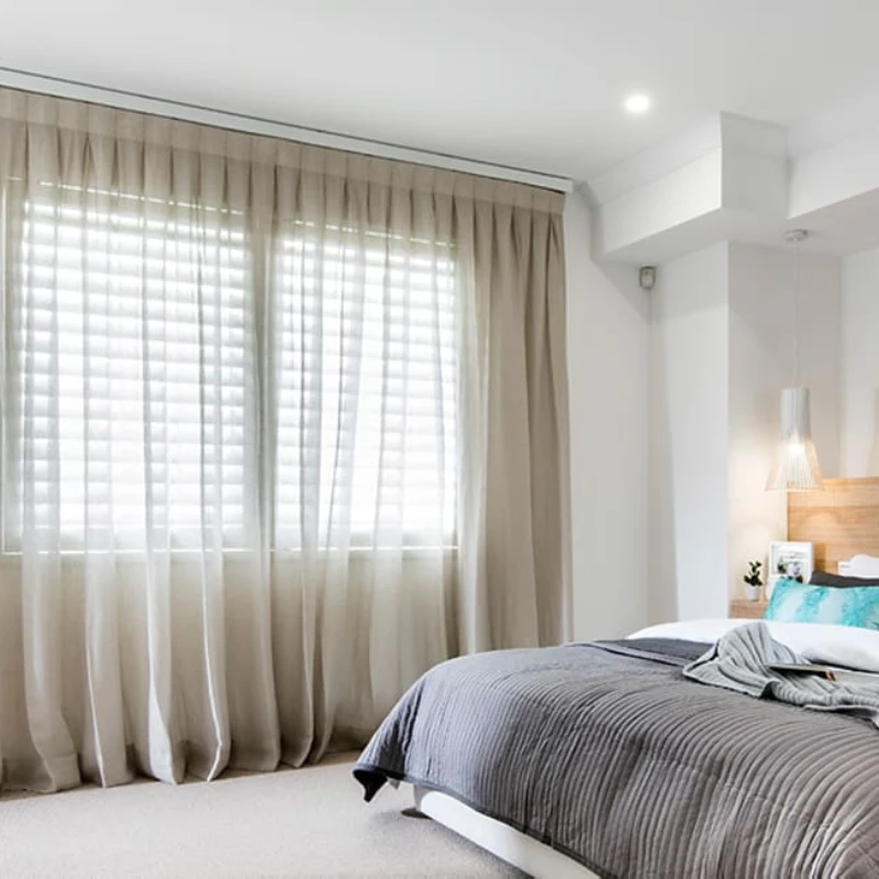 Modern bedroom featuring motorized light beige sheer curtains over white shutters for smart privacy.